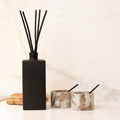 A black reed diffuser bottle with four black reeds and the words 'THE NAUGHTY FORK' printed on the front, styled on a white marble countertop with two marble candle holders in the background.