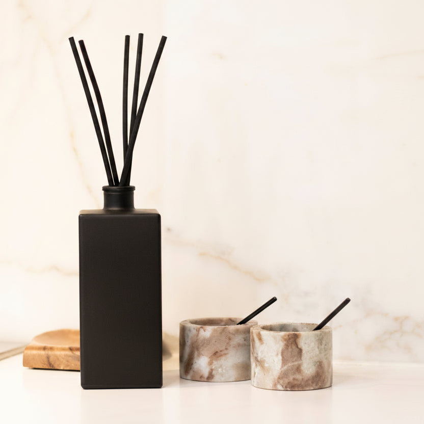 A black reed diffuser bottle with four black reeds and the words 'THE NAUGHTY FORK' printed on the front, styled on a white marble countertop with two marble candle holders in the background.