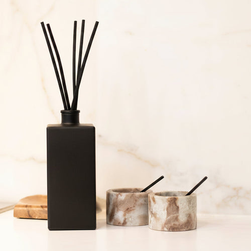 A black reed diffuser bottle with four black reeds and the words 'THE NAUGHTY FORK' printed on the front, styled on a white marble countertop with two marble candle holders in the background.