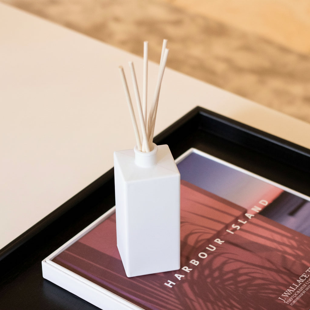 A white rectangular reed diffuser bottle with natural reed sticks, placed on top of a printed magazine or book inside a black tray. The bottle is branded 'THE MATLIN FAMILY'.